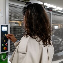 A person operates the control panel of a semi-automatic parking system to retrieve a parked vehicle. In the background, the movable parking spaces and a vehicle within the parking facility are visible.